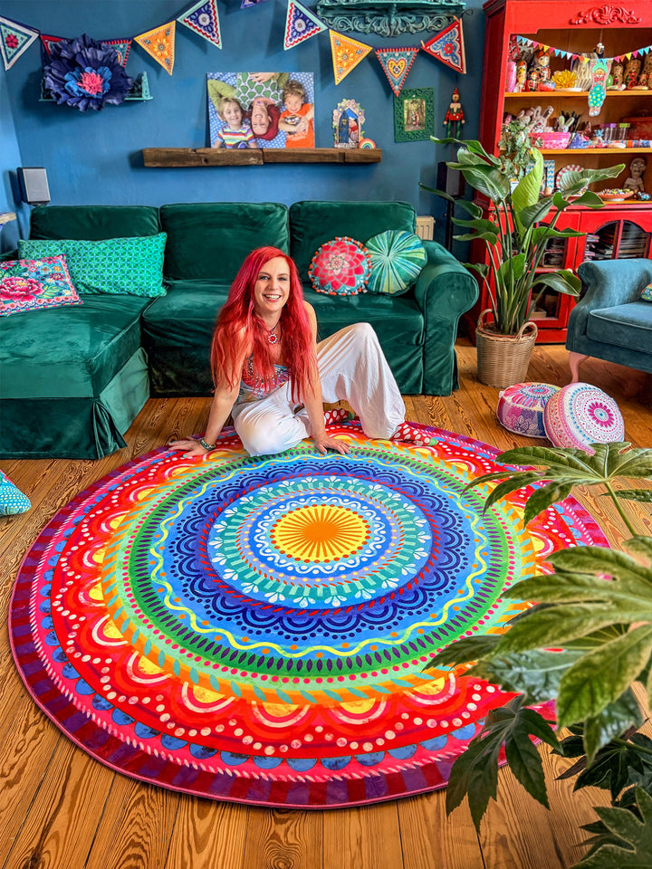 Runder Teppich mit farbenfrohem Mandala-Muster in Blau, Türkis, Gelb, Grün, Pink, Rot und Lila liegt auf warmem Holzboden in einem gemütlichen Wohnzimmer. Im Hintergrund stehen ein großes grünes Samtsofa, Zimmerpflanzen, bunte Kissen und dekorative Möbel vor einer dunkelblauen Wand mit Girlanden und Wandkunst. Der farbige Teppich setzt einen lebendigen Mittelpunkt und schafft eine kreative, wohnliche Boho-Atmosphäre.
