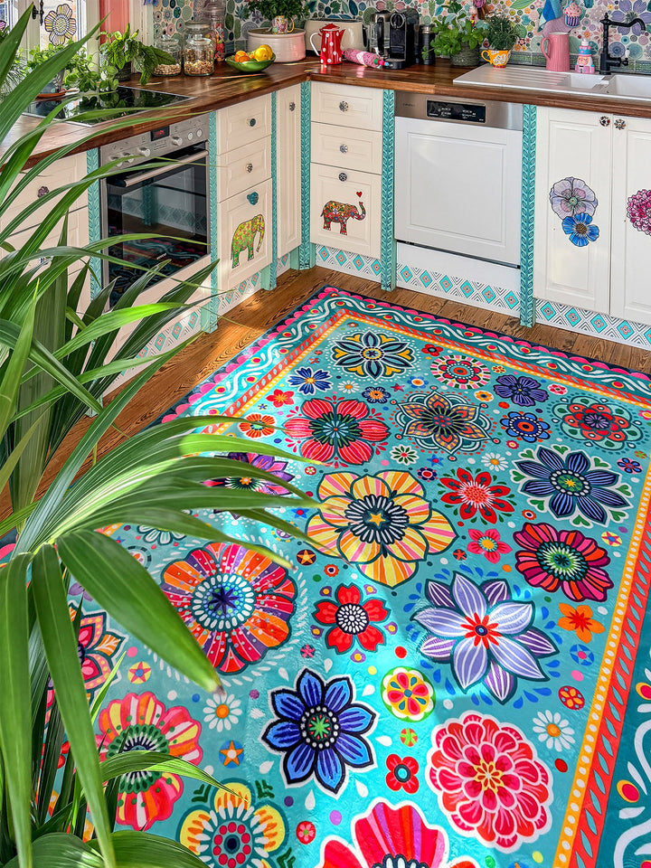 Großer Teppich in Türkis mit farbenfrohen Blumen- und Mandala-Mustern in Pink, Gelb, Rot, Blau, Grün und Lila liegt auf Holzboden in einer kreativ gestalteten Küche. Im Hintergrund stehen helle Küchenschränke mit dekorativen Elefantenmotiven, Holzarbeitsplatten, Pflanzen, Obstschalen und bunte Fensterdekorationen. Der florale Teppich bringt lebendige Farben, gemütliche Wärme und verspielten Boho-Stil in den Küchenbereich.