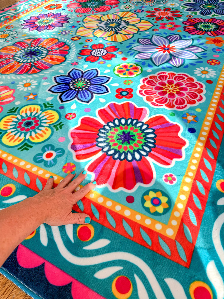 Großer Teppich in Türkis mit farbenfrohen Blumen- und Mandala-Mustern in Pink, Gelb, Rot, Blau, Grün und Lila liegt auf Holzboden in einer kreativ gestalteten Küche. Im Hintergrund stehen helle Küchenschränke mit dekorativen Elefantenmotiven, Holzarbeitsplatten, Pflanzen, Obstschalen und bunte Fensterdekorationen. Der florale Teppich bringt lebendige Farben, gemütliche Wärme und verspielten Boho-Stil in den Küchenbereich.