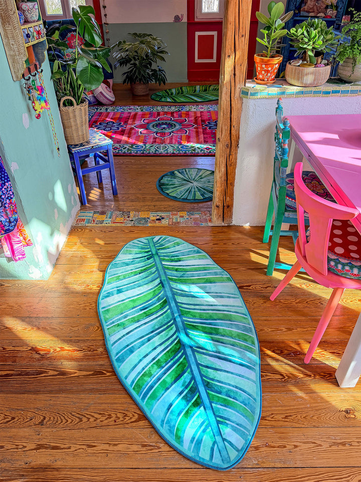 Grüner Blatt-Teppich liegt auf warmem Holzboden in einem farbenfrohen Wohnraum mit kreativer Einrichtung. Im Hintergrund stehen viele Zimmerpflanzen, ein pinker Tisch mit passenden Stühlen sowie weitere bunte Teppiche mit Mandala- und Blumenmustern. Sonnenlicht fällt in den Raum und betont die natürlichen Farben. Die Szene wirkt gemütlich, verspielt und tropisch inspiriert.