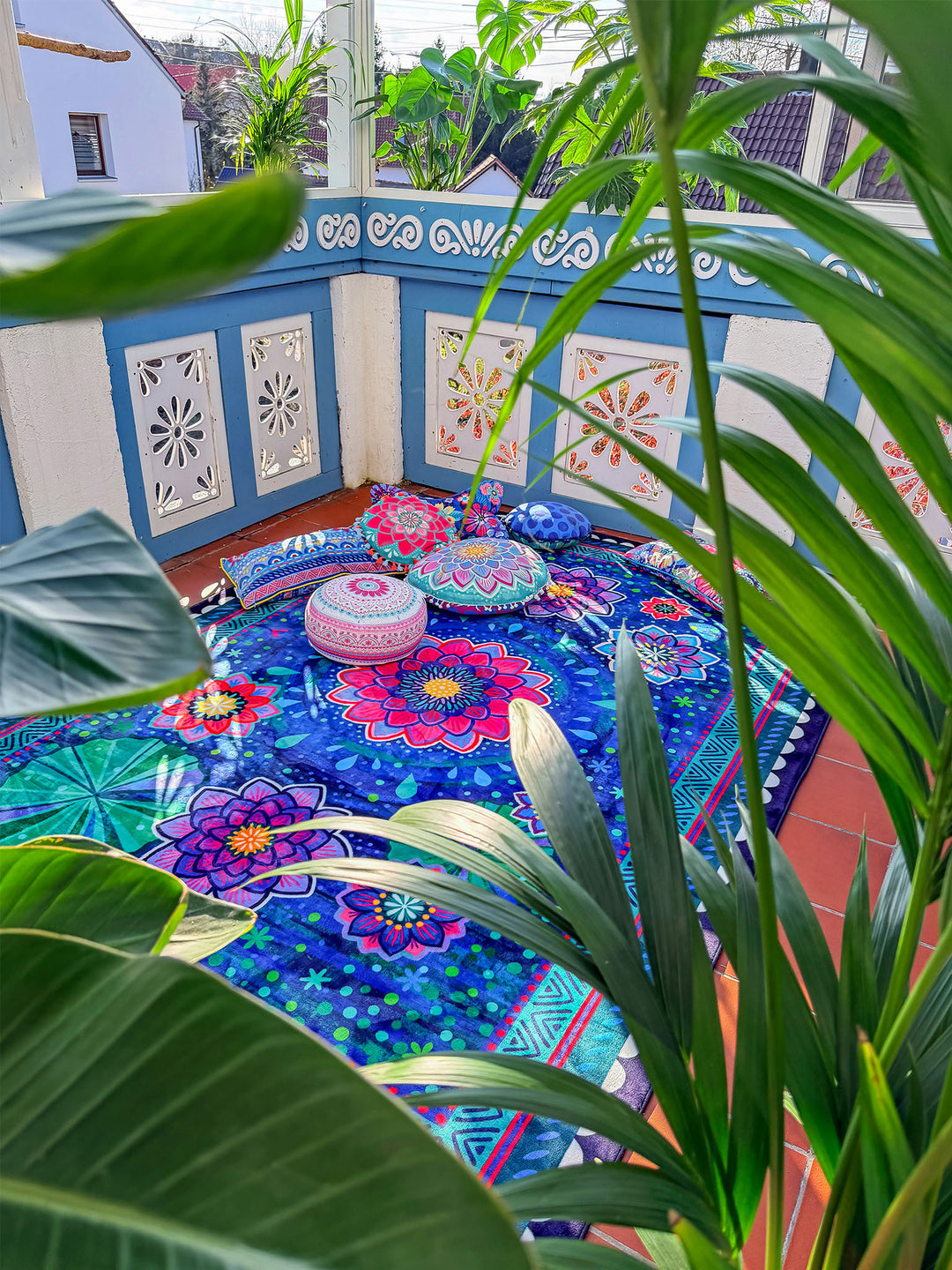 Großer Teppich mit floralem Mandala-Muster in Dunkelblau, Pink, Lila, Türkis und Grün liegt in einem hellen Wintergarten oder auf einer geschützten Terrasse. Auf dem Teppich liegen dekorative Kissen und ein runder Sitzhocker mit bunten Mustern. Im Vordergrund rahmen rosa und weiße Blüten sowie grüne Pflanzen die Szene ein. Sonnenlicht, Pflanzen und farbenfrohe Textilien schaffen eine gemütliche, lebendige Boho-Wohnatmosphäre.