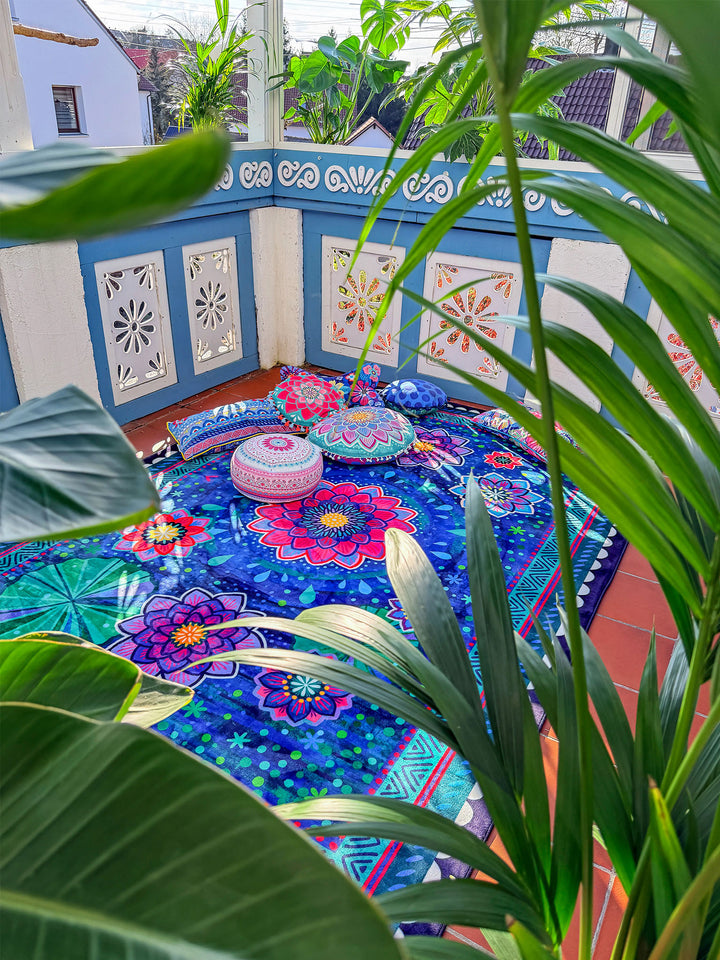 Großer Teppich mit floralem Mandala-Muster in Dunkelblau, Pink, Lila, Türkis und Grün liegt in einem hellen Wintergarten oder auf einer geschützten Terrasse. Auf dem Teppich liegen dekorative Kissen und ein runder Sitzhocker mit bunten Mustern. Im Vordergrund rahmen rosa und weiße Blüten sowie grüne Pflanzen die Szene ein. Sonnenlicht, Pflanzen und farbenfrohe Textilien schaffen eine gemütliche, lebendige Boho-Wohnatmosphäre.