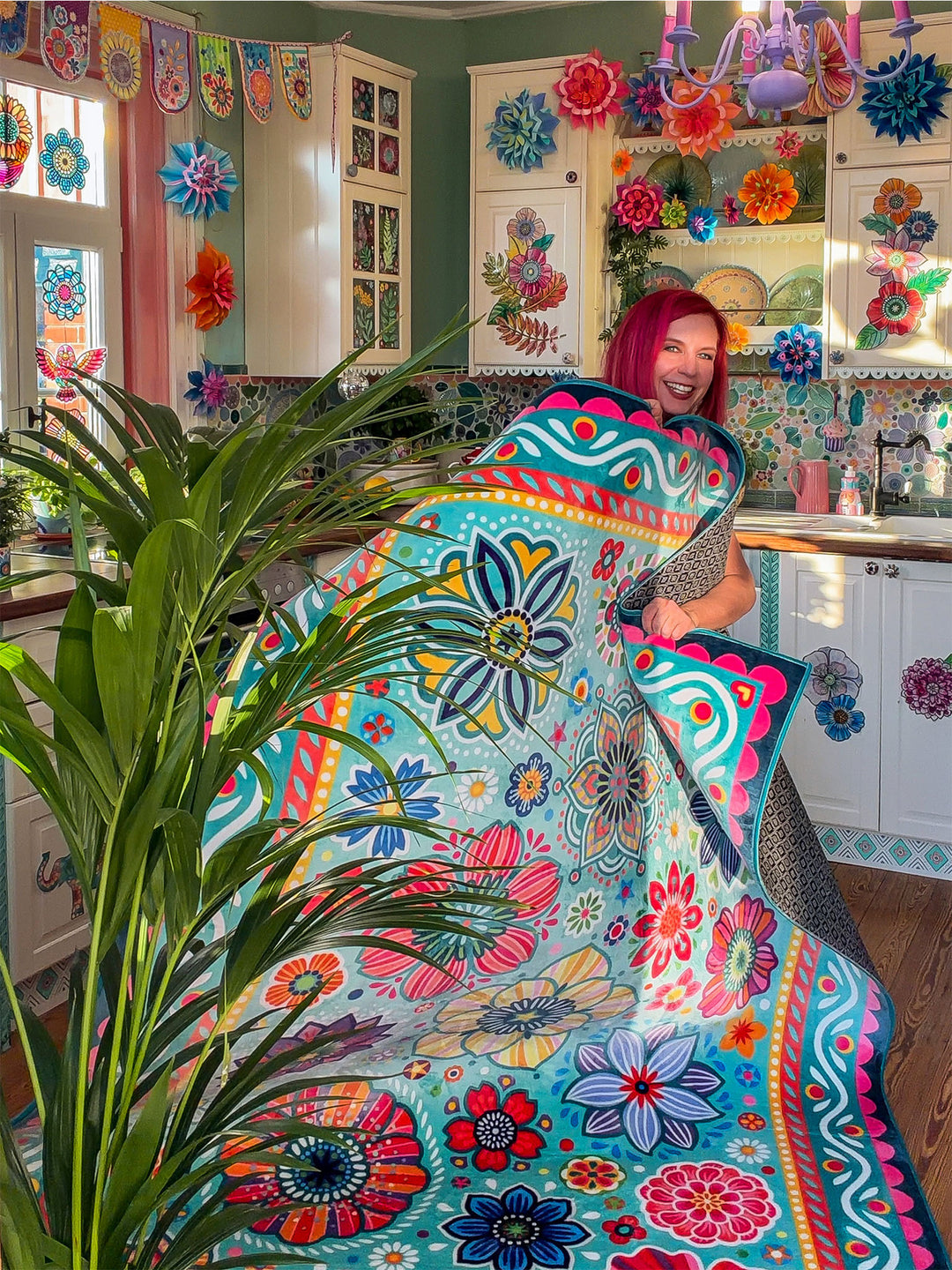 Bine Brändle steht lächelnd in einer farbenfroh dekorierten Küche und hält einen großen Teppich mit Blumen- und Ornamentmustern hoch. Der Teppich ist türkisfarben und mit bunten Details in Pink, Gelb, Rot, Blau und Grün gestaltet. Im Vordergrund steht eine große Zimmerpflanze mit langen grünen Blättern. Die kreative Küche ist mit bunten Papierblumen, dekorierten Schränken und Mosaikfliesen geschmückt. Die Szene wirkt lebendig, verspielt und voller fröhlicher Farben.