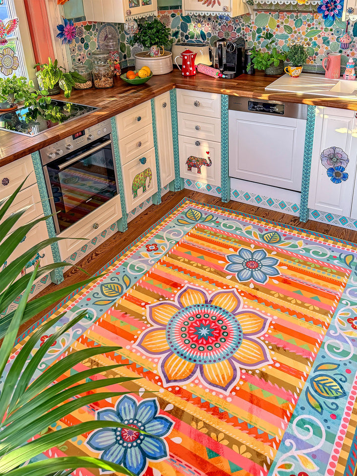 Großer Küchenteppich mit farbenfrohem Blumen- und Ornamentmuster in Orange, Gelb, Türkis, Blau, Rosa und Grün liegt auf Holzboden vor einer hellen Landhausküche. Im Hintergrund stehen weiße Küchenschränke mit dekorativen Elefantenmotiven, Holzarbeitsplatte, Backofen und Geschirrspüler. Der bunte Teppich bringt Wärme, kreative Farben und gemütlichen Boho-Stil in den Küchenbereich.
