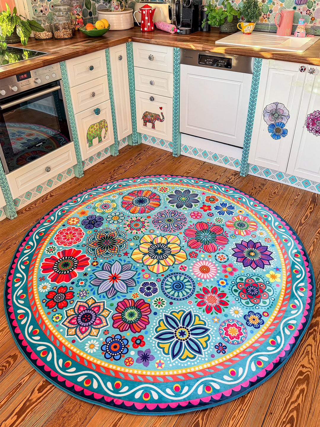Runder Teppich in Türkis mit farbenfrohen Blumen- und Mandala-Mustern in Pink, Gelb, Rot, Blau, Grün und Lila liegt auf warmem Holzboden in einer kreativ gestalteten Küche. Im Hintergrund stehen helle Küchenschränke mit dekorativen Elefanten- und Blumenmotiven, Holzarbeitsplatten, Pflanzen sowie moderne Küchengeräte. Der bunte Teppich setzt einen lebendigen Mittelpunkt und bringt gemütlichen Boho-Stil in den Küchenbereich.