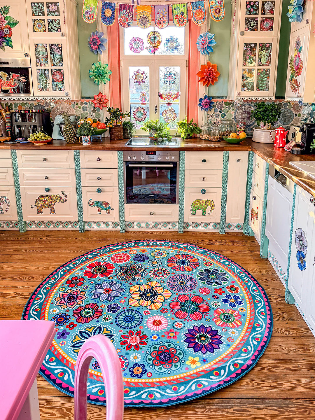 Runder Teppich in Türkis mit farbenfrohen Blumen- und Mandala-Mustern in Pink, Gelb, Rot, Blau, Grün und Lila liegt auf warmem Holzboden in einer kreativ gestalteten Küche. Im Hintergrund stehen helle Küchenschränke mit dekorativen Elefanten- und Blumenmotiven, Holzarbeitsplatten, Pflanzen sowie moderne Küchengeräte. Der bunte Teppich setzt einen lebendigen Mittelpunkt und bringt gemütlichen Boho-Stil in den Küchenbereich.