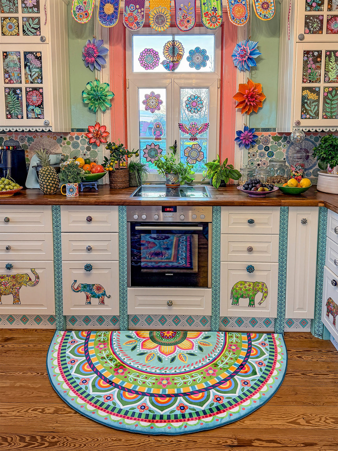 Halbrunder Küchenteppich in Pastell-Türkis mit detailreichen Mandala- und Blumenmustern in Pink, Gelb, Grün, Orange und Blau liegt vor einer hellen Landhausküche mit Holzarbeitsplatte. Die weißen Schränke sind mit farbenfrohen Elefantenmotiven verziert. Über dem Spülbecken befindet sich ein dekoriertes Fenster mit bunten Blumenstickern und Papierblüten. Pflanzen, Obstschalen und liebevolle Details schaffen eine warme, kreative Boho-Atmosphäre.
