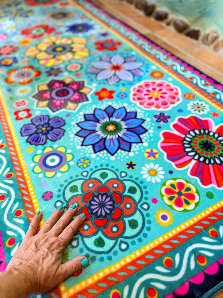 Schmaler rechteckiger Badteppich in Türkis mit farbenfrohen Blumen- und Mandalamotiven in Pink, Blau, Gelb, Rot und Grün liegt auf terrakottafarbenen Fliesen vor einer Badewanne. Das kreative Badezimmer ist mit kunstvollen Mosaikwänden, Naturstein-Details und Pflanzen dekoriert. Große grüne Blätter und ein Fenster mit Tageslicht schaffen eine frische, natürliche Atmosphäre. Der lebendige Teppich setzt einen fröhlichen Farbakzent im individuell gestalteten Boho-Bad.