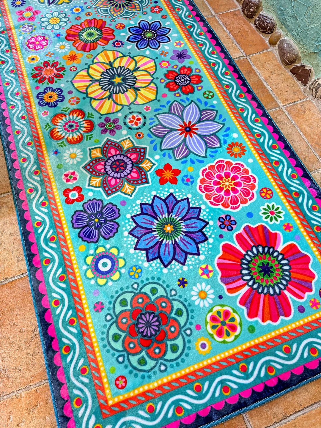 Schmaler rechteckiger Badteppich in Türkis mit farbenfrohen Blumen- und Mandalamotiven in Pink, Blau, Gelb, Rot und Grün liegt auf terrakottafarbenen Fliesen vor einer Badewanne. Das kreative Badezimmer ist mit kunstvollen Mosaikwänden, Naturstein-Details und Pflanzen dekoriert. Große grüne Blätter und ein Fenster mit Tageslicht schaffen eine frische, natürliche Atmosphäre. Der lebendige Teppich setzt einen fröhlichen Farbakzent im individuell gestalteten Boho-Bad.