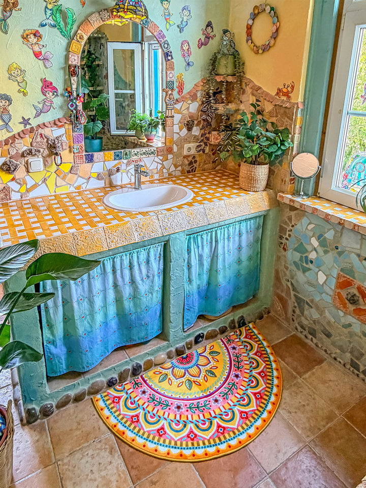 Halbrunder Badteppich in leuchtendem Gelb mit farbenfrohem Mandala- und Blumenmuster in Pink, Türkis, Orange, Blau und Grün liegt auf warmen Natursteinfliesen vor einem Waschtisch. Der rustikal-kreative Waschtisch besitzt eine gelb-weiß geflieste Ablage, ein weißes Waschbecken und türkisfarbene Stoffvorhänge darunter. Naturstein- und Mosaikdetails an Wand und Möbeln verleihen dem Badezimmer einen kunstvollen Boho-Stil. Die fröhliche Matte setzt einen lebendigen Farbakzent im Raum.
