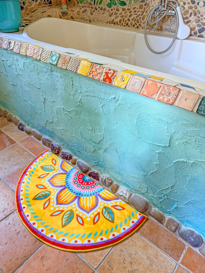 Halbrunder Badteppich in kräftigem Gelb mit großem Blütenmandala in Pink, Türkis, Lila, Orange und Grün liegt auf terrakottafarbenen Fliesen vor einer Badewanne. Die Wanne ist von einer türkis verputzten Front mit Natursteinabschluss und einer bunten Bordüre aus handgefertigten Fliesen eingefasst. Im Hintergrund sorgen Mosaiksteinwände und natürliche Materialien für eine mediterrane Boho-Atmosphäre. Die farbenfrohe Matte setzt einen fröhlichen Blickfang im individuell gestalteten Badezimmer.