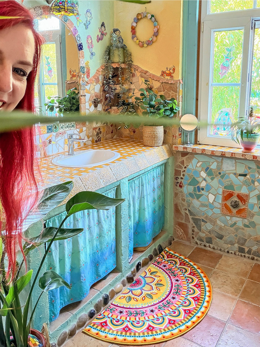 Halbrunder Badteppich in leuchtendem Gelb mit farbenfrohem Mandala- und Blumenmuster in Pink, Türkis, Orange, Blau und Grün liegt auf warmen Natursteinfliesen vor einem Waschtisch. Der rustikal-kreative Waschtisch besitzt eine gelb-weiß geflieste Ablage, ein weißes Waschbecken und türkisfarbene Stoffvorhänge darunter. Naturstein- und Mosaikdetails an Wand und Möbeln verleihen dem Badezimmer einen kunstvollen Boho-Stil. Die fröhliche Matte setzt einen lebendigen Farbakzent im Raum.
