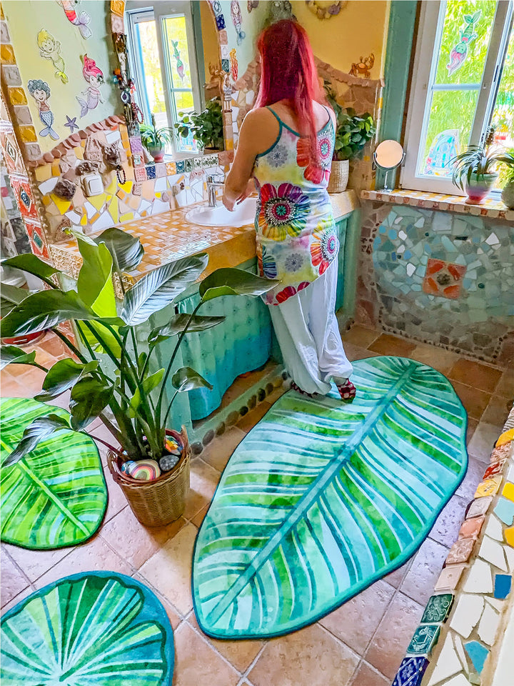 Künstlerisch gestaltetes Badezimmer im Boho- und Tropenstil mit mehreren grünen Badteppichen in Blattform auf terrakottafarbenen Fliesen. Die Teppiche erinnern an große tropische Blätter mit detailreichen Blattadern in verschiedenen Grün- und Türkistönen. Im Mittelpunkt steht eine große Zimmerpflanze im Korb, umgeben von den Teppichen. Der Waschtisch ist mit gelb-weißen Mosaikfliesen verkleidet und besitzt türkisfarbene Stoffvorhänge als Stauraum. Wände, Fensterrahmen und Badewanne sind mit Naturstein, bunt