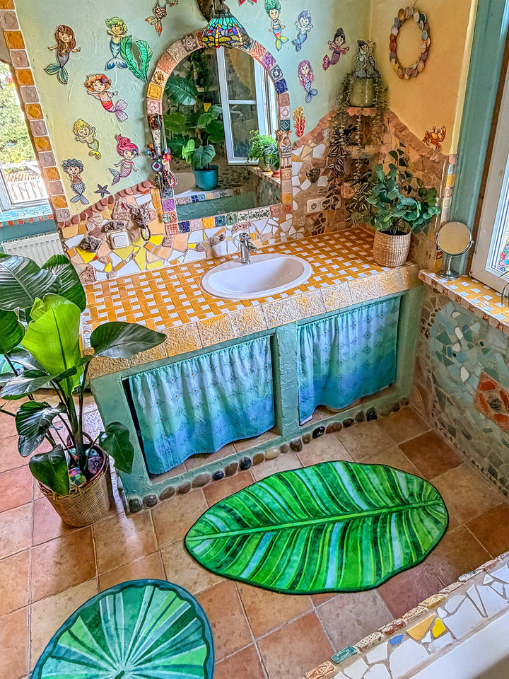 Farbenfrohes Badezimmer im Boho-Stil mit mehreren dekorativen Badteppichen auf warmen Terrakottafliesen. Im Vordergrund liegt ein großer Teppich in Form eines tropischen Blattes mit detailreichen Blattadern in satten Grün- und Limetönen. Dahinter ergänzen ein runder grüner Blätterteppich und ein halbrunder Mandala-Teppich in Türkis, Pink, Gelb und Blau das harmonische Gesamtbild. Links steht eine große Zimmerpflanze im geflochtenen Korb, rechts befindet sich eine Badewanne mit türkis verputzter Front und bu