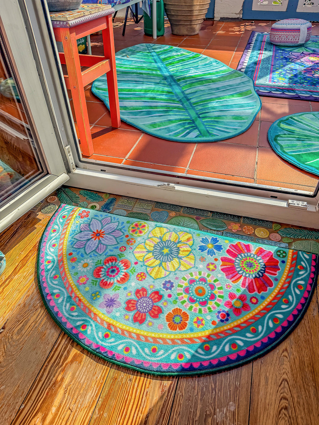 Halbrunder Teppich in Türkis liegt vor einer geöffneten Terrassentür auf warmem Holzboden. Der Teppich ist mit bunten Blumen, Mandala-Ornamenten und verspielten Mustern in Pink, Gelb, Rot, Orange, Blau und Grün gestaltet. Im Außenbereich auf roten Fliesen liegen grüne Blatt-Teppiche. Die helle Szene verbindet farbenfrohes Wohndesign mit natürlicher, sommerlicher Wohnatmosphäre.