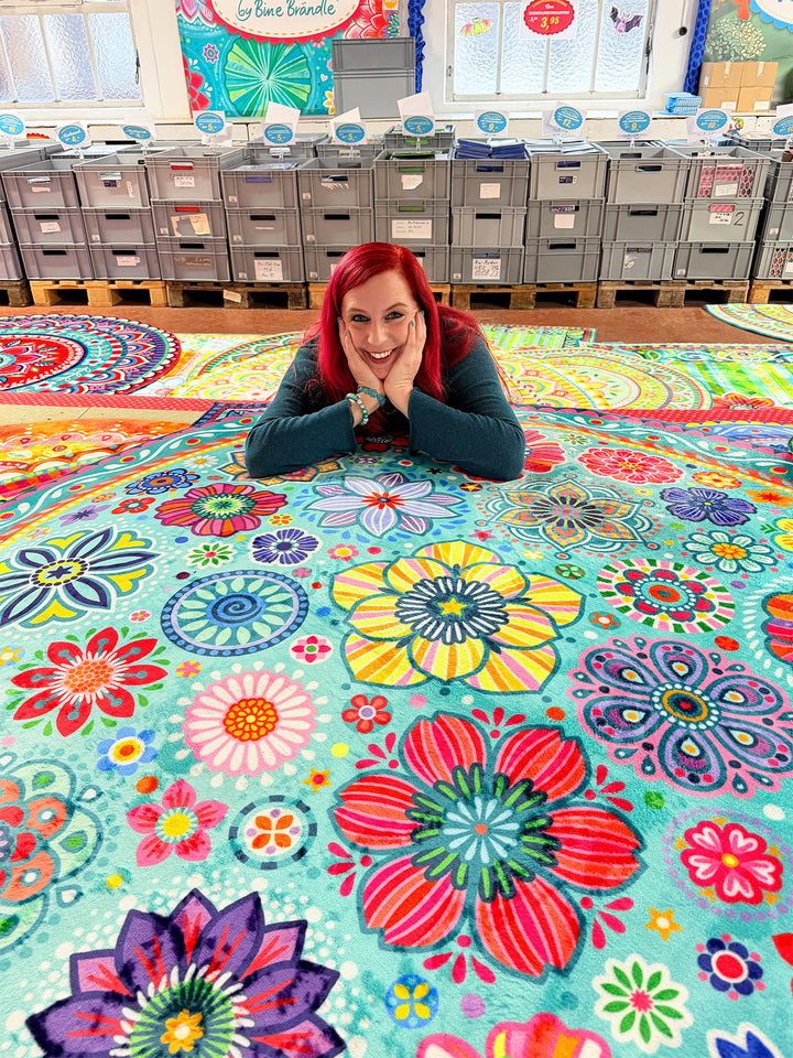 Bine Brändle liegt lächelnd hinter einem großen türkisfarbenen Teppich mit bunten Blumen, Ornamenten und Mandala-Mustern in Pink, Gelb, Rot, Blau und Grün. Im Hintergrund stehen Lagerboxen und dekorierte Wände eines Showrooms.