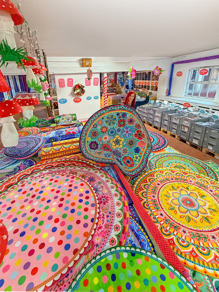 Innenaufnahme eines bunt dekorierten Verkaufsraums. Im Vordergrund liegen viele große, runde Teppiche mit farbenfrohen Mustern, Blumenornamenten und Punkten in Pink, Grün, Gelb, Blau und Orange übereinander gestapelt. In der Mitte hält eine Person einen weiteren runden Teppich hoch und lächelt in die Kamera. Links hängen große dekorative Pilzfiguren von der Decke. Rechts stehen Reihen grauer Kisten auf Paletten. Im Hintergrund sind weitere Warenregale, Fenster und bunte Dekorationen zu sehen. Die Szene wirk